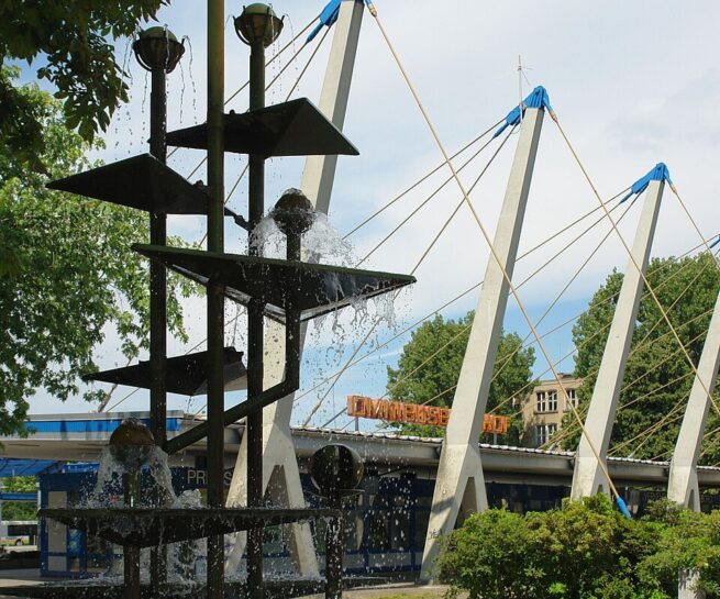 abstrakte Skulptur im Vordergrund. Masten eines Gebäudes im Hintergrund